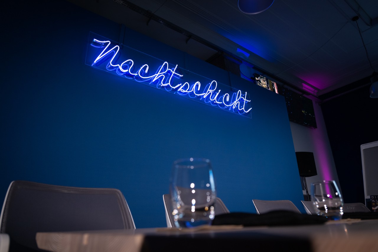 Ein leerer Tisch, auf dem dem Gläser stehen. Dahinter leuchtet an der Wand in blauer Farbe der Name der Veranstaltung: „Nachtschicht“.