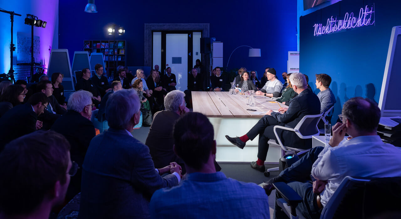 In front of an audience, several people sit at a table in front of a dark blue wall with the illuminated lettering Nachtschicht.