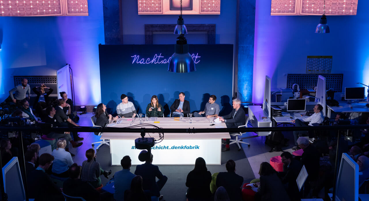 In front of an audience, six people sit at a large table in front of a dark blue wall with a neon sign.