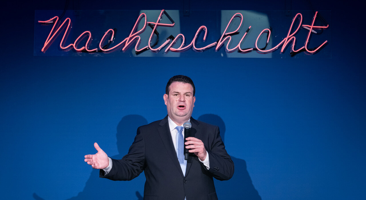 Hubertus Heil speaks into a microphone and stands in front of a dark blue wall with the illuminated lettering Nachtschicht.