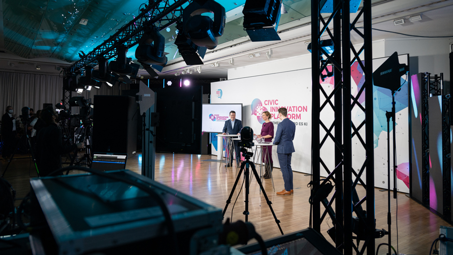 Drei Personen stehen auf einer Bühne an Stehtischen neben einem Monitor und vor einer Rückwand mit der Aufschrift Civic Innovation Platform.
