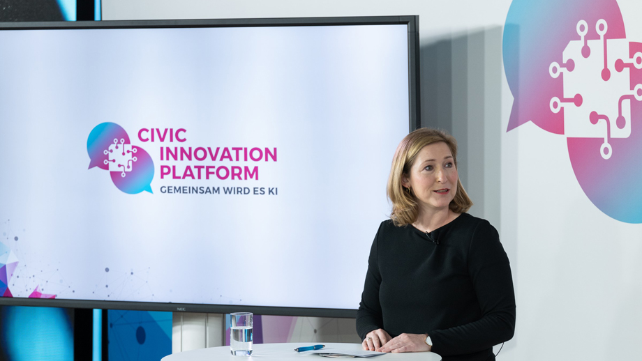 Eine Frau steht an einem Stehtisch neben einem großen Monitor auf dem das Logo der Civic Innovation Platform zu sehen ist.