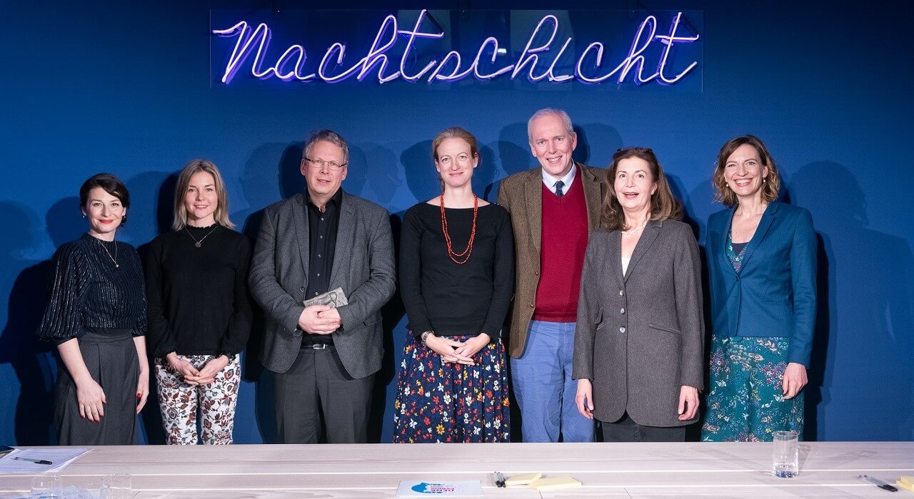 Five women and two men stand in front of a dark blue wall with the illuminated lettering Nachtschicht.