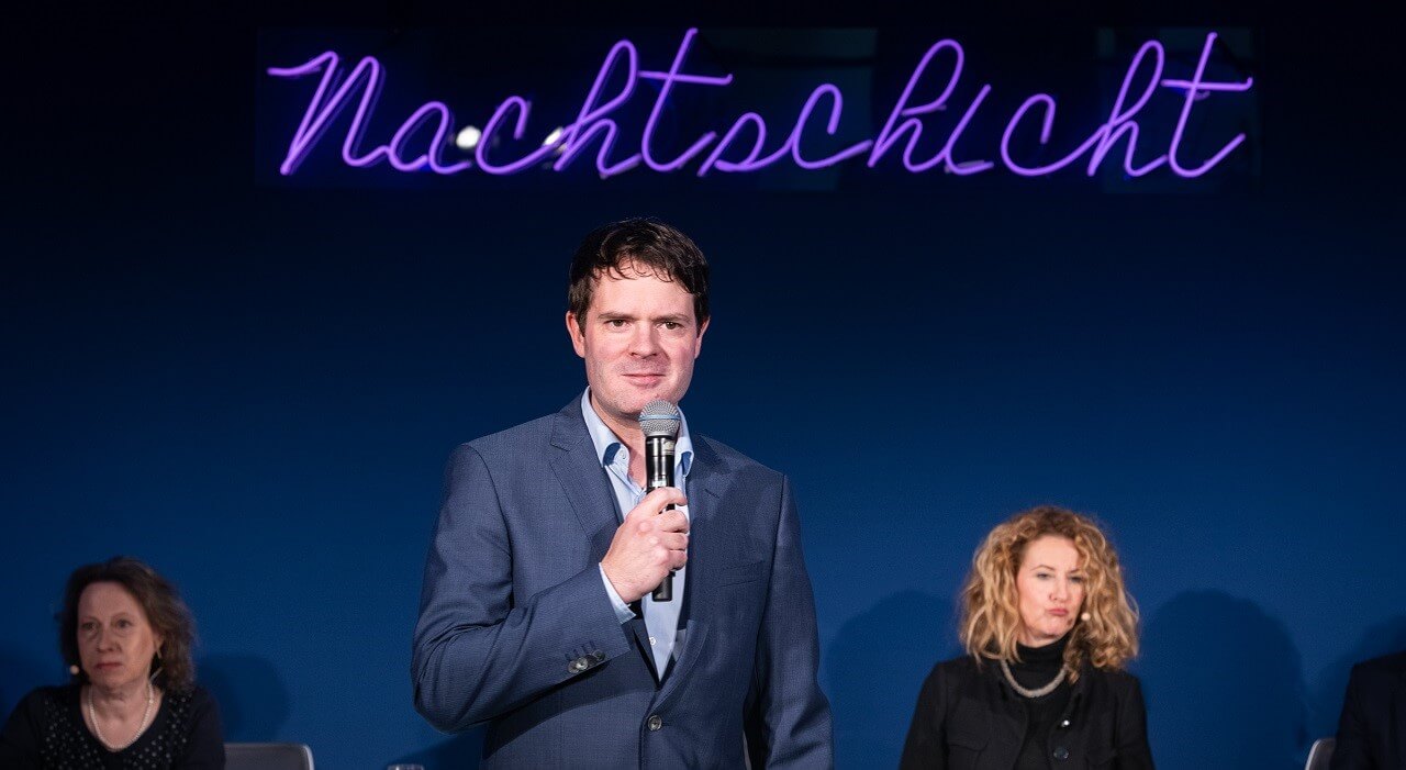 A man in a suit with a microphone stands in front of a dark blue wall with the illuminated lettering Nachtschicht.