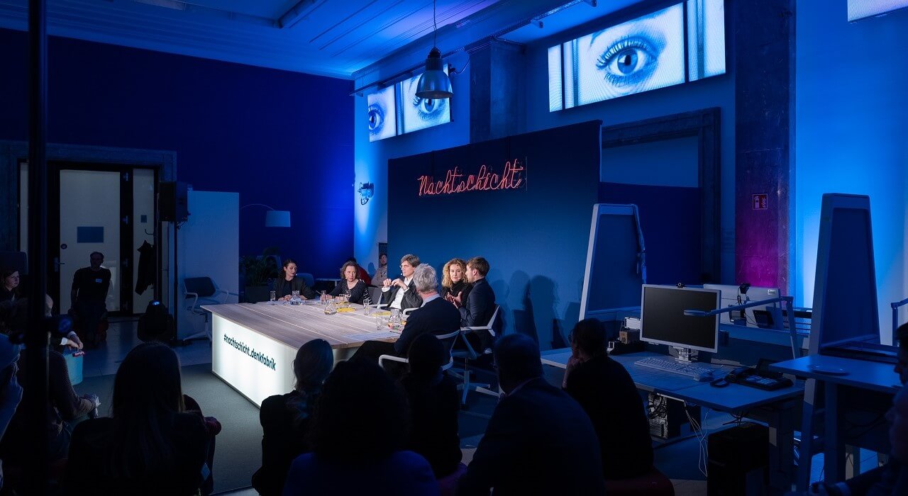 Several people sit in front of an audience at a table in front of a back wall with the illuminated lettering Nachtschicht.
