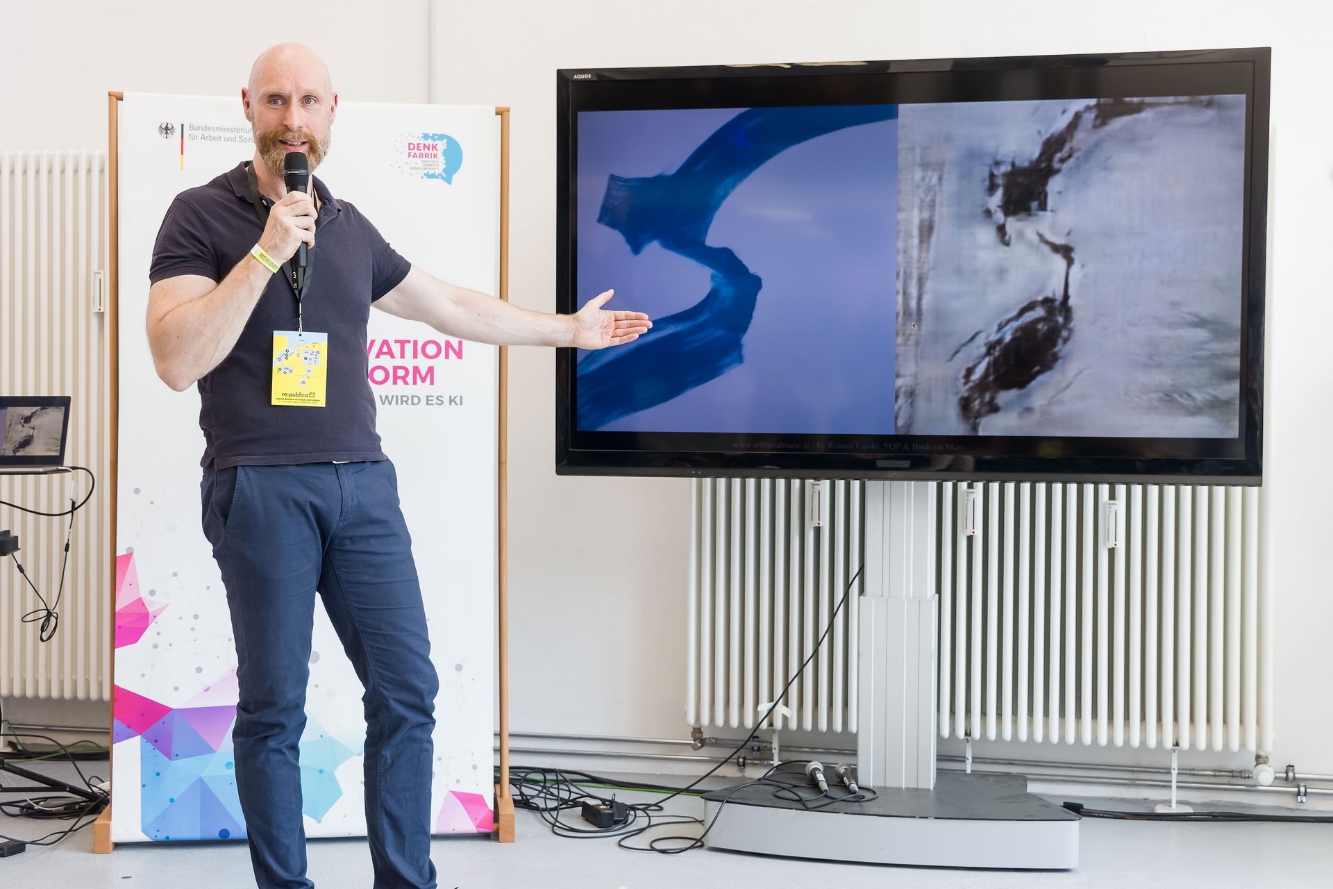 Der Künstler Roman Lipski zeigt auf seine Bilder, welche auf einem Bildschirm gezeigt werden. Er hält ein Mikrofon in der Hand und steht vor einem Plakat der Denkfabrik.