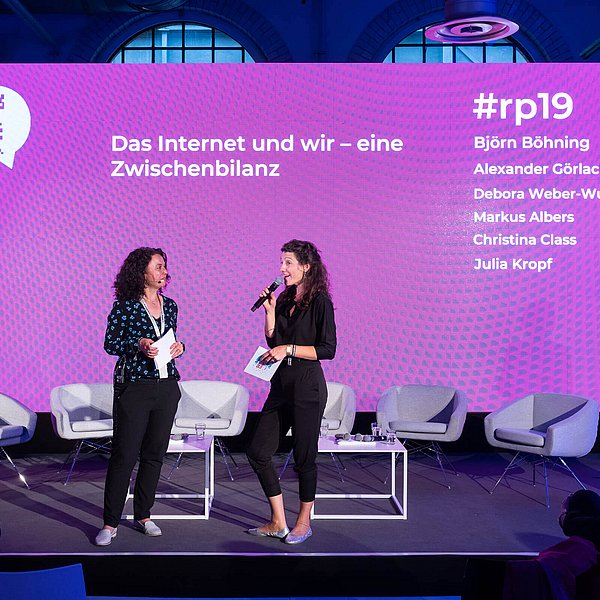 Zwei Frauen mit Mikrofonen und Karten in der Hand stehen auf einer Bühne vor einer magentafarbenen Rückwand mit dem Titel Das Internet und wir - eine Zwischenbilanz.