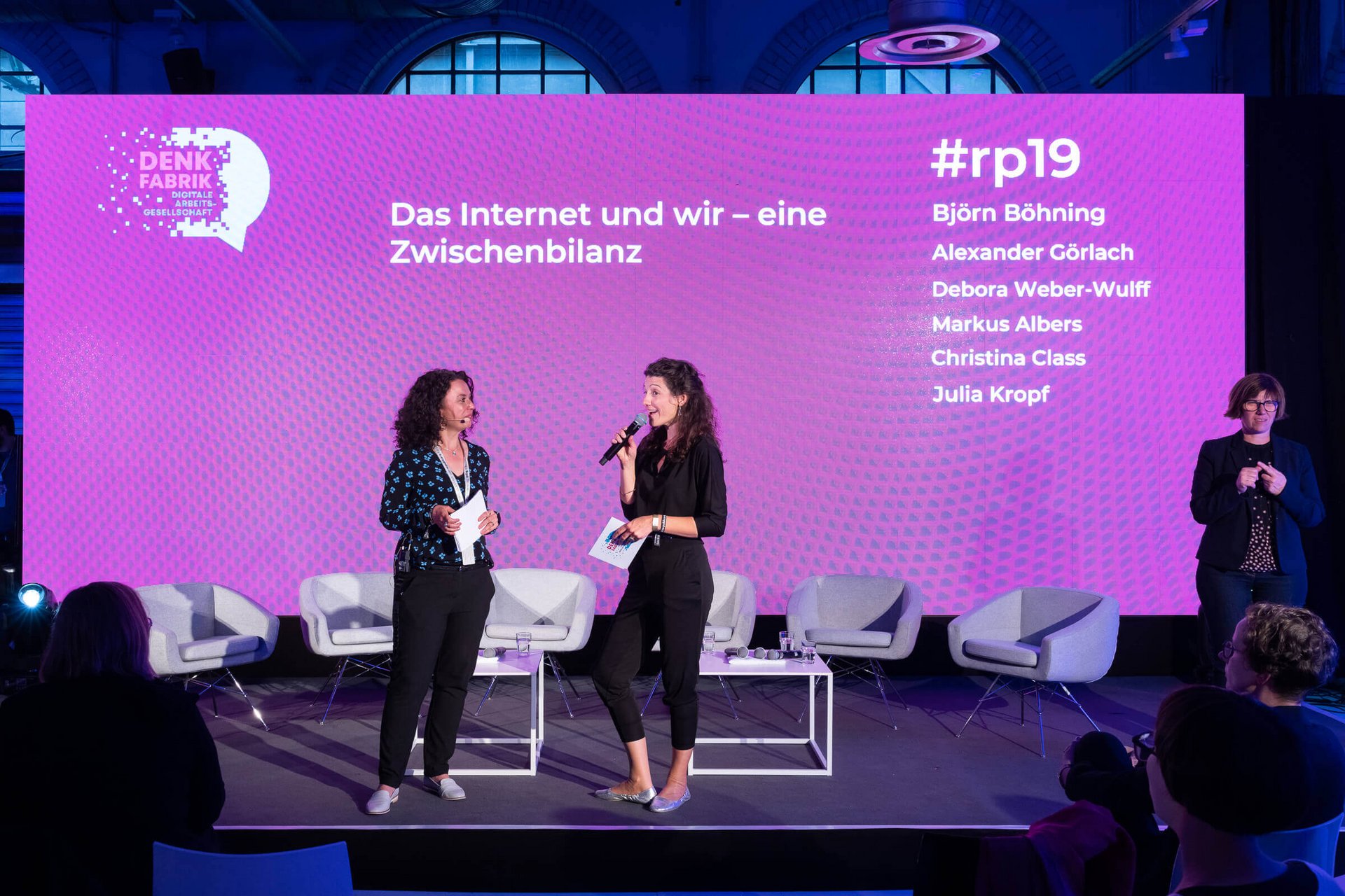 Zwei Frauen mit Mikrofonen und Karten in der Hand stehen auf einer Bühne vor einer magentafarbenen Rückwand mit dem Titel Das Internet und wir - eine Zwischenbilanz.