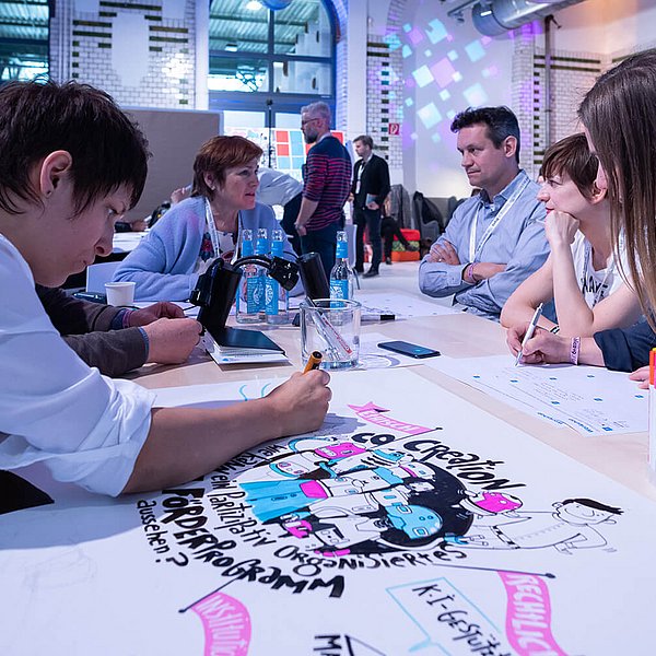Eine Frau sitzt mit mehreren Personen an einem Tisch und zeichnet etwas auf ein bereits mit Illustrationen und Text bestücktes Bild.