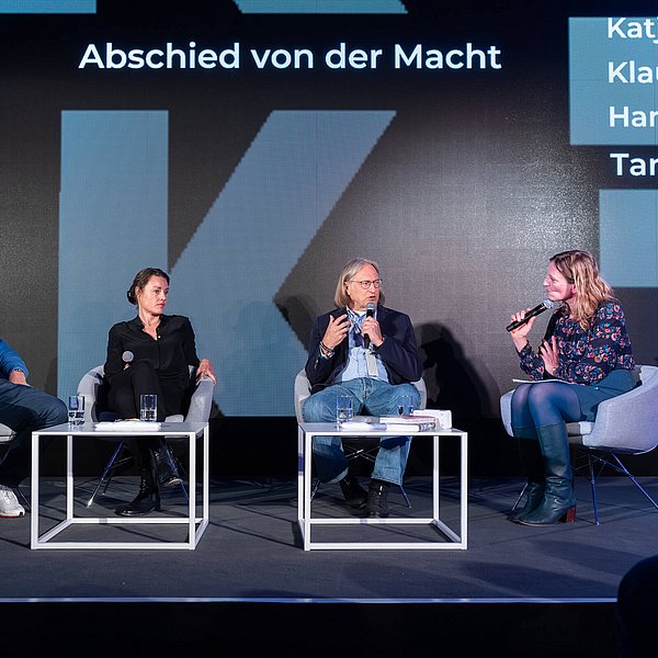 Vier Personen sitzen auf einer Bühne vor einer Rückwand mit der Aufschrift Abschied von der Macht. Zwei Personen davon sprechen in ein Mikrofon.