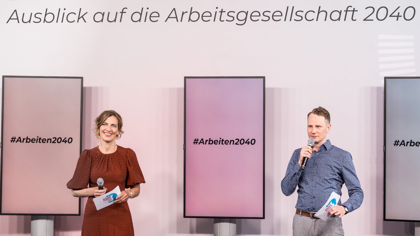 Zwei Personen mit Mikrofonen und Karten in der Hand stehen auf einer Bühne vor Monitoren mit der Aufschrift #Arbeiten2040.