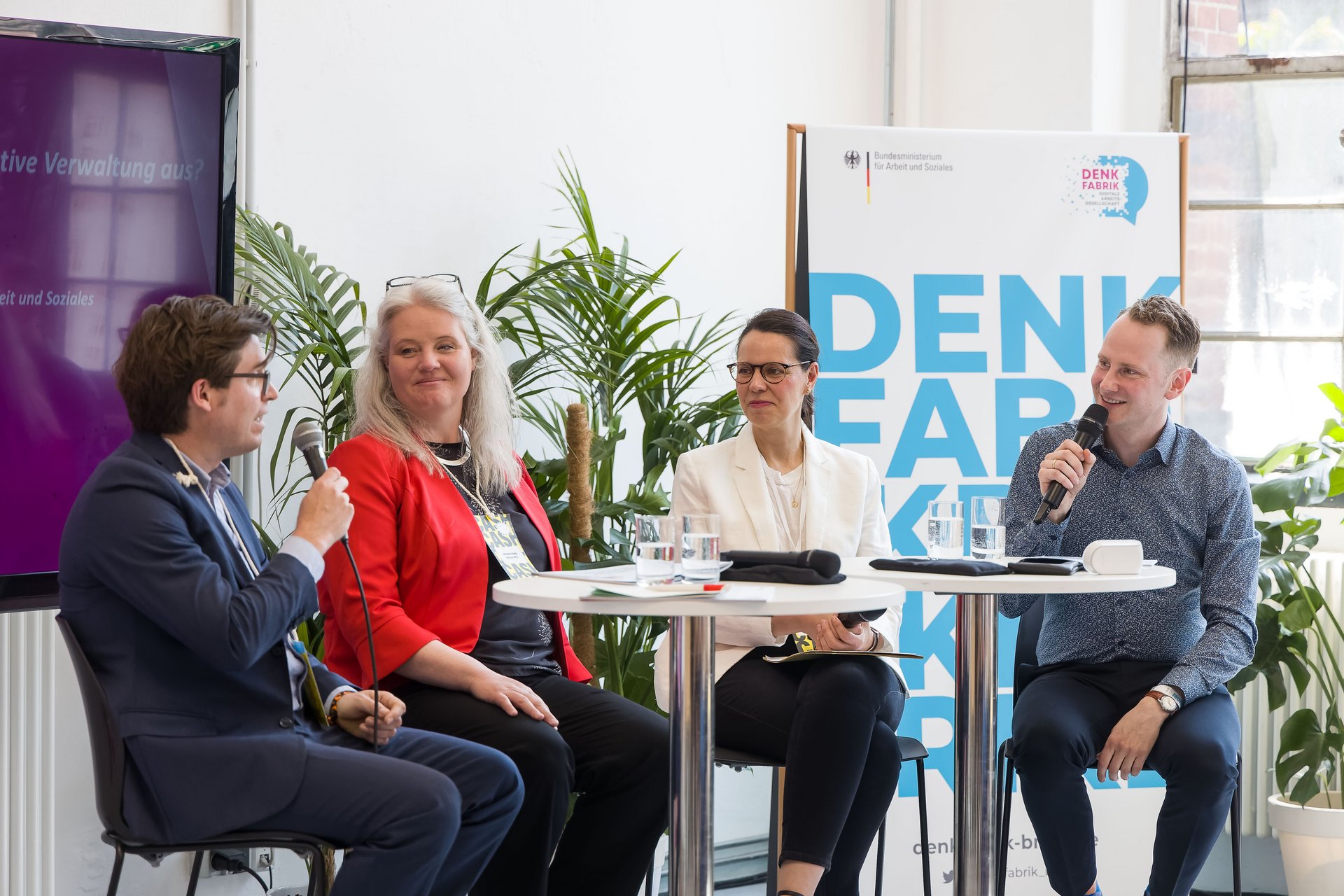 Jakob Kort, stellvertretender Leiter der Abteilung Digitalisierung und Unternehmensentwicklung, spricht mit Ana Dujíc, Abteilungsleiterin der Denkfabrik, Professorin Katharina Zweig und Moderator Mads Pankow. Im Hintergrund sind ein Plakat der Denkfabrik und Pflanzen.