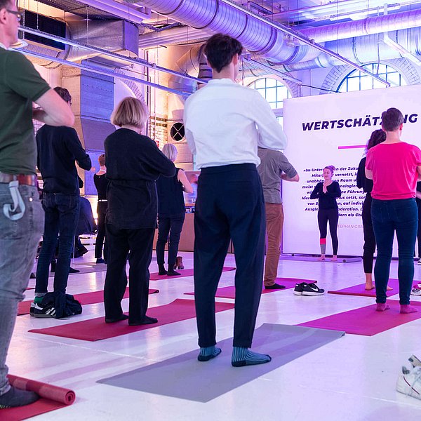 Mehrere Personen stehen in einer Industriehalle auf Gymnastkmatten einer Frau gegenüber und machen Yoga-Übungen.