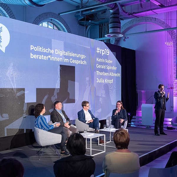 Vier Personen sitzen und eine Person steht auf einer Bühne vor einer Rückwand mit dem Titel Politische Digitalisierungsberater*innen im Gespräch.