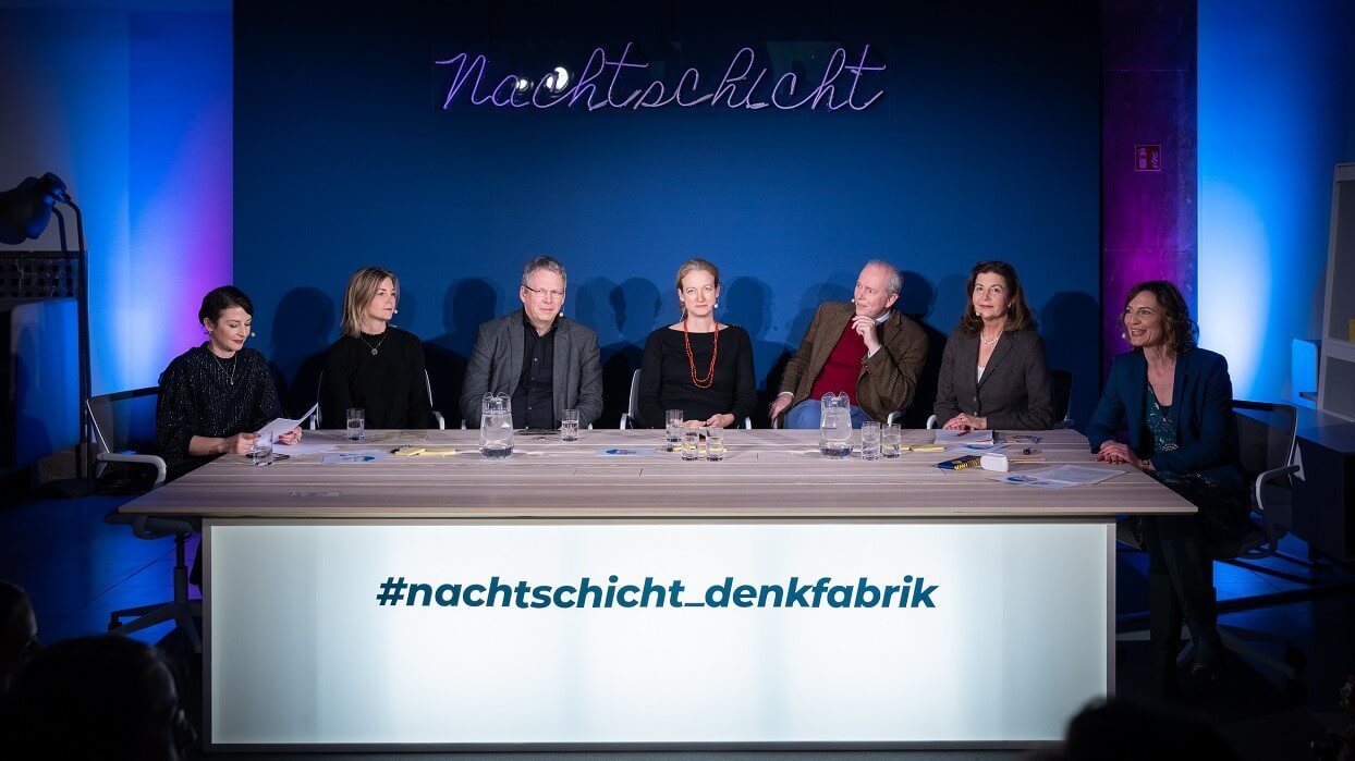 Sieben Personen sitzen an einem Tisch mit der Aufschrift #nachtschicht-denkfabrik vor einer dunkelblauen Rückwand mit dem Schriftzug Nachtschicht.