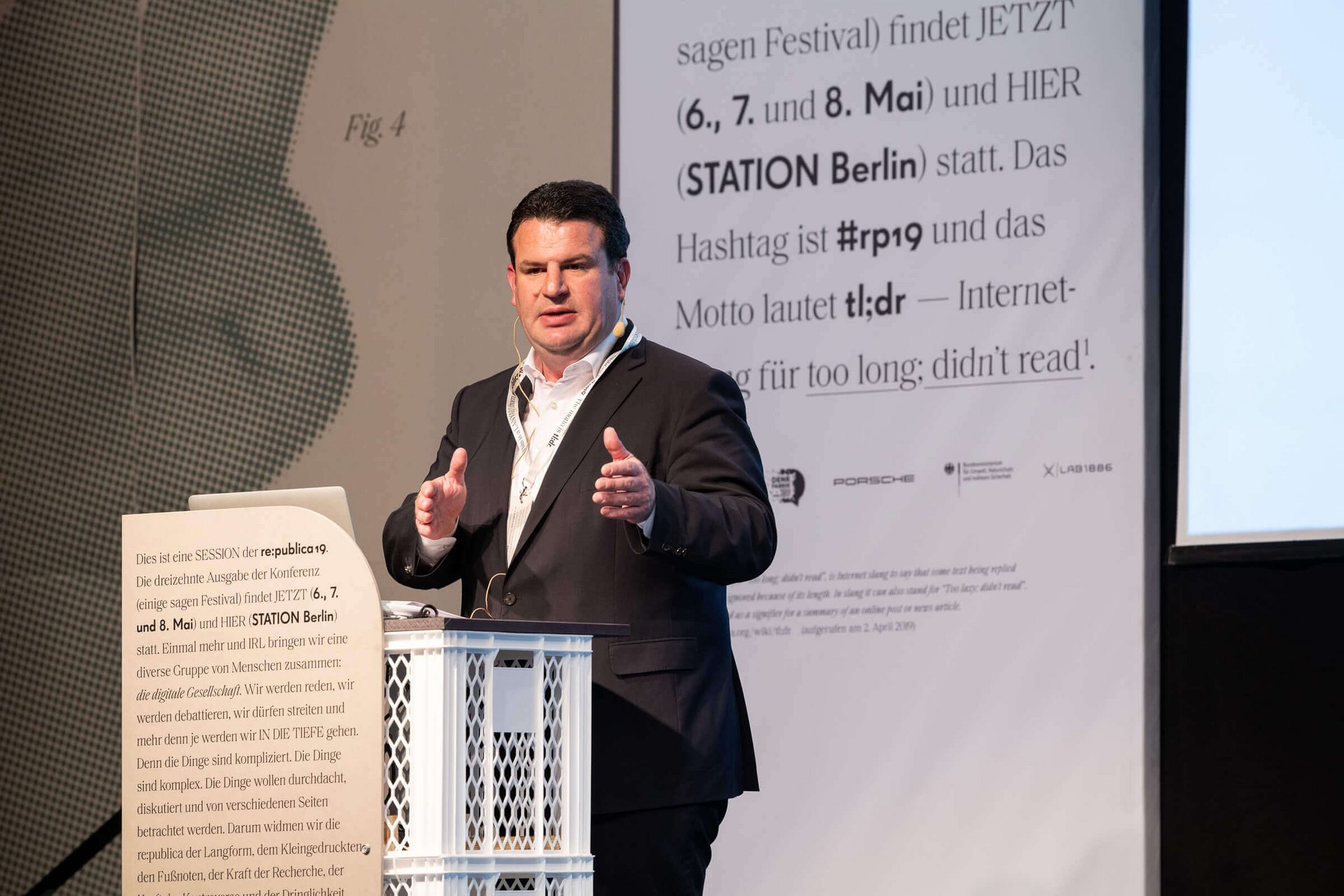 Hubertus Heil trägt ein Kopfbügelmikrofon, steht auf einer Bühne und spricht.