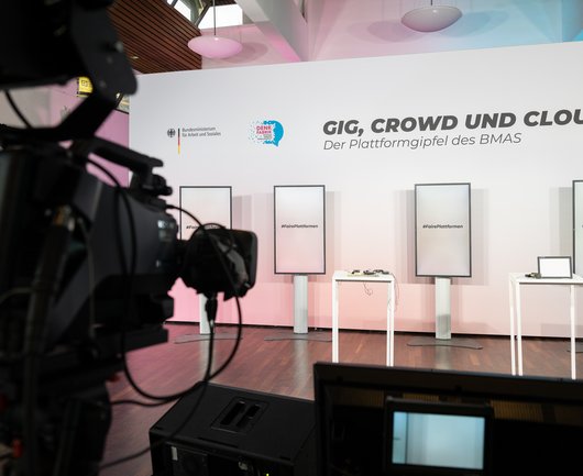 Eine Kamera ist auf eine Bühne mit vier Monitoren und zwei Tischen gerichtet. Auf der Bühnenrückwand steht Gig, Crowd, Cloud. Der Plattformgipfel des BMAS. 